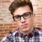 Portrait of a young man wearing eyeglasses and a plaid shirt against a brick wall.